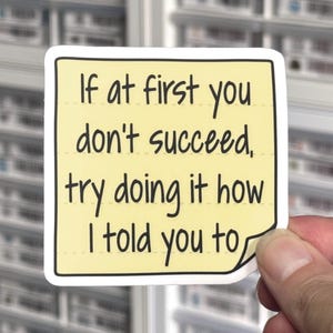 May include: A square yellow adhesive note sticker with rounded corners and a curled bottom right edge. The sticker features black handwritten-style text that reads: If at first you don't succeed, try doing it how I told you to.