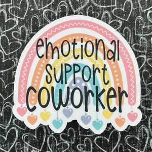 Puede incluir: Una pegatina troquelada con un arco&iacute;ris de colores pastel y el texto negro "emotional SUPPORT COWORKER" impreso en el centro. El arco&iacute;ris tiene peque&ntilde;os dijes en forma de coraz&oacute;n colgando de su base en tonos rosa, amarillo y azul.