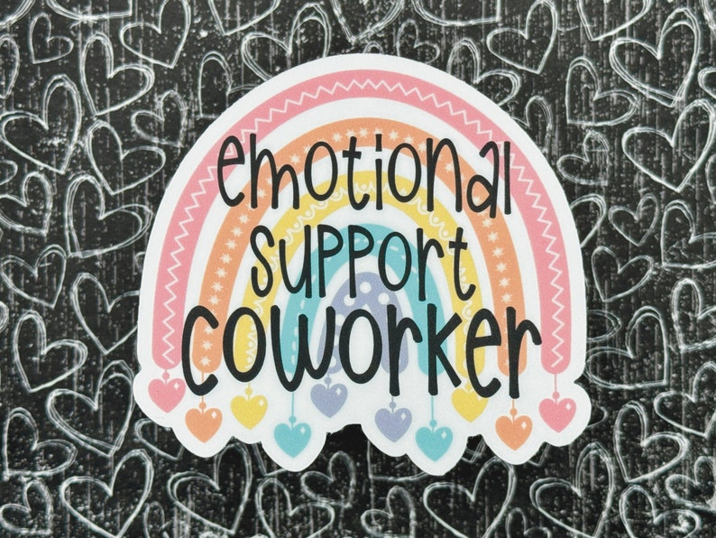 Puede incluir: Una pegatina troquelada con un arco&iacute;ris de colores pastel y el texto negro "emotional SUPPORT COWORKER" impreso en el centro. El arco&iacute;ris tiene peque&ntilde;os dijes en forma de coraz&oacute;n colgando de su base en tonos rosa, amarillo y azul.