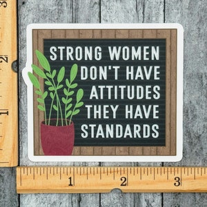 May include: A square sticker featuring a black letter board with a wood-look frame and the white text "STRONG WOMEN DON'T HAVE ATTITUDES THEY HAVE STANDARDS." A potted green plant sits to the left. A wooden ruler shows the sticker is 3 inches wide.