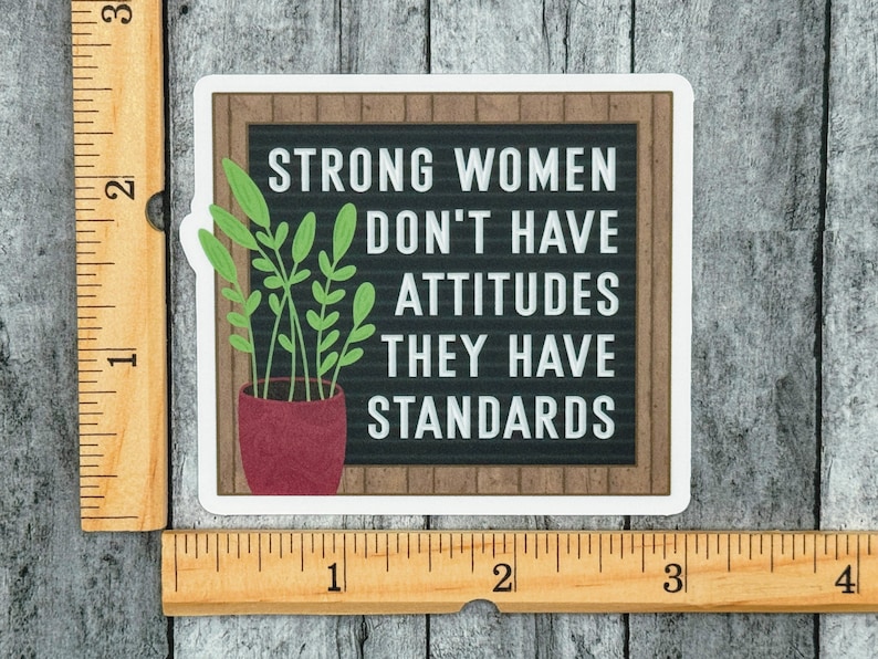 May include: A square sticker featuring a black letter board with a wood-look frame and the white text "STRONG WOMEN DON'T HAVE ATTITUDES THEY HAVE STANDARDS." A potted green plant sits to the left. A wooden ruler shows the sticker is 3 inches wide.