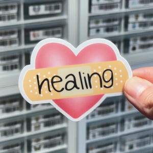 May include: A heart-shaped sticker featuring a pink gradient background with a beige adhesive bandage graphic placed horizontally across the center. The word healing is printed in black, lowercase, handwritten-style lettering on the bandage.