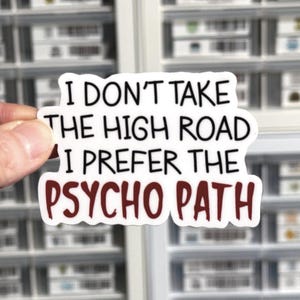 May include: A die-cut vinyl sticker with a white background and black text that reads I DON'T TAKE THE HIGH ROAD I PREFER THE, followed by the word PSYCHO PATH in bold, dark red, hand-lettered style font, featuring a clean, irregular border.