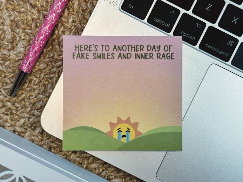 Pu&ograve; includere: Un post-it rosa e giallo con il testo "HERE'S TO ANOTHER DAY OF FAKE SMILES AND INNER RAGE". Un sole dei cartoni animati con un viso triste e lacrime &egrave; in fondo al post-it.