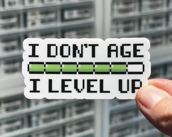 May include: A rectangular white sticker with black pixelated text that reads I DON'T AGE I LEVEL UP. Between the two lines of text is a horizontal progress bar with a black outline, filled with segments of light green color, indicating progress.