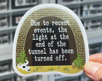 May include: A die-cut sticker featuring a brick tunnel entrance with a dark interior. The text reads: Due to recent events, the light at the end of the tunnel has been turned off. A white animal skull and green desert plants sit on the yellow ground.