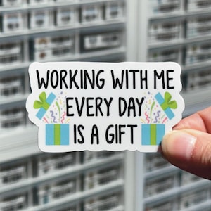 May include: A white sticker with the text "WORKING WITH ME EVERY DAY IS A GIFT" in black. The sticker features two open gift boxes with blue and green accents, and colorful confetti.