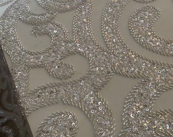 May include: White fabric with a delicate pattern of silver sequins and beads. The fabric is designed in a repeating pattern of swirls and curves. The fabric is perfect for a wedding dress or other formal wear.
