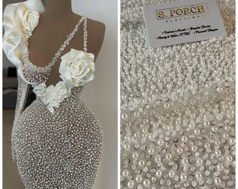 May include: A white dress made of pearl beads with a white satin flower detail. The dress is on a mannequin. The fabric is shown in a close-up image. The fabric is made of pearl beads and is very delicate. The fabric is perfect for a special occasion dress.