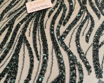 May include: Close-up of a fabric with a repeating pattern of dark green sequins and beads. The sequins are in a wave-like design, with a sheer, light-coloured background. A small white tag with text is visible in the upper left corner.