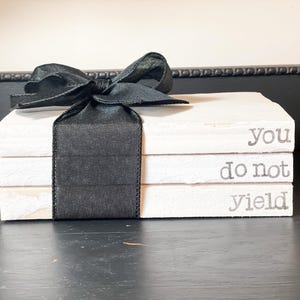 Puede incluir: Una pila de tres bloques de madera blancos atados con una cinta negra. Los bloques tienen las palabras "You do not yield" impresas en gris. La cinta está atada con un lazo.