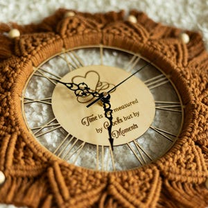 May include: A decorative wall clock with a macrame design in shades of brown. The clock face is made of wood with Roman numerals and the quote "Time is measured by clocks but by Moments". The clock has a diameter of approximately 30 cm.