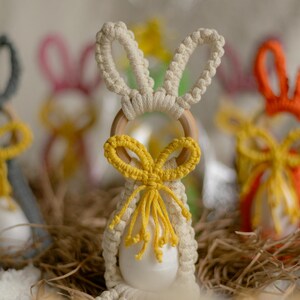 May include: Macrame bunny decorations in cream, grey, pink, yellow, and orange. Each bunny has a wooden ring and a decorative bow. The bunnies are arranged on straw, creating an Easter theme. The bunnies are approximately 10 cm tall.