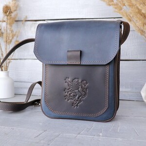 May include: A blue and brown leather crossbody bag with a flap closure and adjustable strap. The front features a detailed embossed lion crest. The bag has a rectangular shape and is accented with brown stitching.