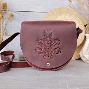 Puede incluir: Bolso bandolera de cuero burdeos con un diseño floral en relieve en la parte delantera. El bolso tiene forma redondeada, una correa larga ajustable y un cierre plateado. Un sombrero de paja y un jarrón decorativo en el fondo.
