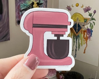 May include: A die-cut sticker featuring a pink stand mixer with a dark gray mixing bowl. The appliance has a solid pink body with a horizontal dark gray accent line across the head. The small text "created.by.ej" is printed at the bottom edge.