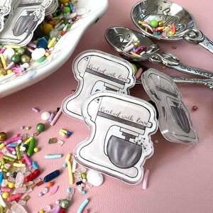 May include: Two clear acrylic stickers with a white stand mixer and the text "Baked with love" in black script. The stickers are on a pink surface with colorful sprinkles scattered around them.