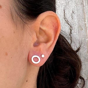 Può includere: Primo piano di un orecchio con due orecchini d'argento. Uno è un piccolo cerchio, l'altro un piccolo punto. Gli orecchini hanno un design semplice e minimalista.