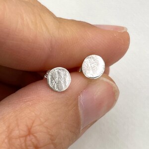 Puede incluir: Un par de pendientes de botón redondos de plata. Cada pendiente tiene una superficie texturizada y se sostiene entre los dedos sobre un fondo blanco. Los pendientes tienen un diseño simple y minimalista.