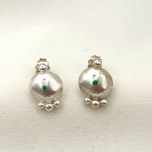 Può includere: Un paio di orecchini a lobo argentati. Ogni orecchino presenta un design arrotondato e bulboso con una piccola pietra verde al centro e tre sfere più piccole sotto. Gli orecchini sono su uno sfondo bianco strutturato.