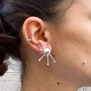 Può includere: Orecchini a lobo in argento dal design unico. Ogni orecchino presenta una sfera centrale argentata con un piccolo accento rosso, da cui si irradiano tre aste argentate. Gli orecchini sono indossati sull'orecchio.