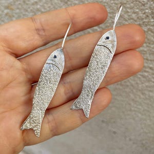 Può includere: Un paio di orecchini a forma di pesce in argento. Ogni orecchino ha un corpo strutturato, una piccola pietra blu scuro per l'occhio e un semplice gancio. Gli orecchini sono progettati per dondolare e catturare la luce.