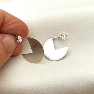 Può includere: Un paio di orecchini a bottone color argento. Ogni orecchino ha una forma a mezzaluna con un ritaglio geometrico. Gli orecchini sono presentati su uno sfondo bianco.