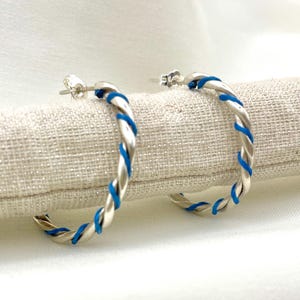 Può includere: Un paio di orecchini a cerchio argentati con un design intrecciato, accentuati da dettagli blu brillante. Gli orecchini sono esposti su una superficie chiara e strutturata, mettendo in risalto la loro finitura lucida e lo stile moderno.