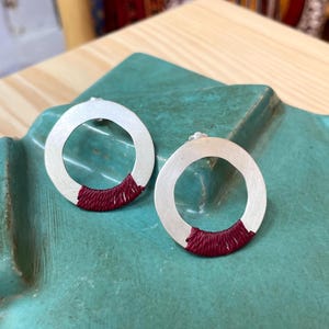 Può includere: Un paio di orecchini a cerchio argentati. Ogni orecchino presenta un design circolare con una sezione di filo rosso scuro avvolta nella parte inferiore. Gli orecchini sono esposti su una superficie color turchese.