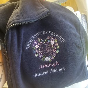 May include: A navy blue fleece jacket with a full-length zip and a high collar. The chest features an embroidered design of a foetus surrounded by a floral heart, with the text "UNIVERSITY OF SALFORD", "Ashleigh", and "Student Midwife".