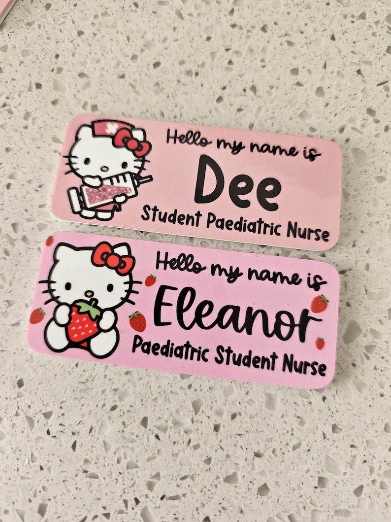 May include: Two rectangular pink name badges featuring Hello Kitty graphics. The top badge reads Hello my name is Dee Student Paediatric Nurse with a syringe graphic. The bottom badge reads Hello my name is Eleanor Paediatric Student Nurse with strawberries.