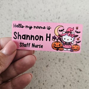 May include: A rectangular pink name tag featuring the black text Hello my name is Shannon H Staff Nurse. The design includes a graphic of a white cat character dressed as a witch with a pink hat, a crescent moon, bats, and orange pumpkins.