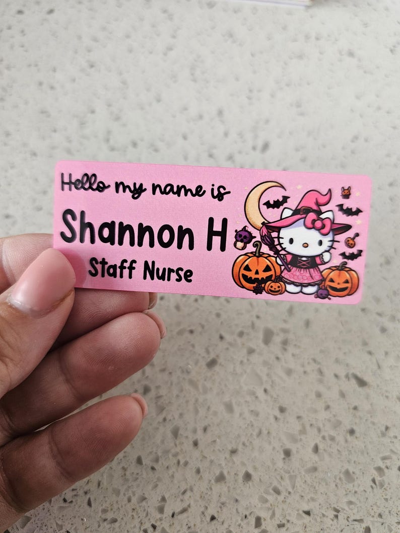 May include: A rectangular pink name tag featuring the black text Hello my name is Shannon H Staff Nurse. The design includes a graphic of a white cat character dressed as a witch with a pink hat, a crescent moon, bats, and orange pumpkins.
