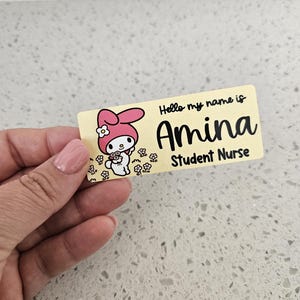 May include: A rectangular name badge with a light yellow background featuring a cartoon character with pink ears holding flowers. The text reads Hello my name is Amina Student Nurse in black lettering. The badge has rounded corners and a smooth finish.