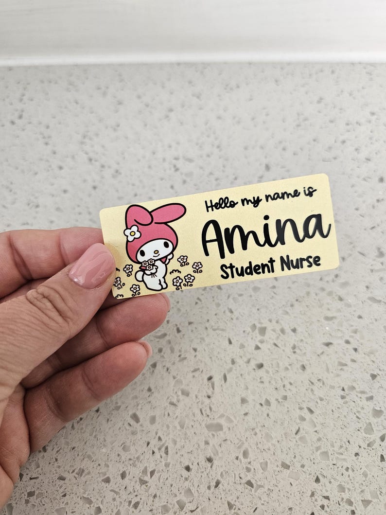 May include: A rectangular name badge with a light yellow background featuring a cartoon character with pink ears holding flowers. The text reads Hello my name is Amina Student Nurse in black lettering. The badge has rounded corners and a smooth finish.