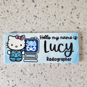 May include: A rectangular name badge with a light blue background featuring a cartoon cat wearing blue scrubs next to an X-ray machine. The text reads Hello my name is Lucy Radiographer in black script and sans-serif fonts.