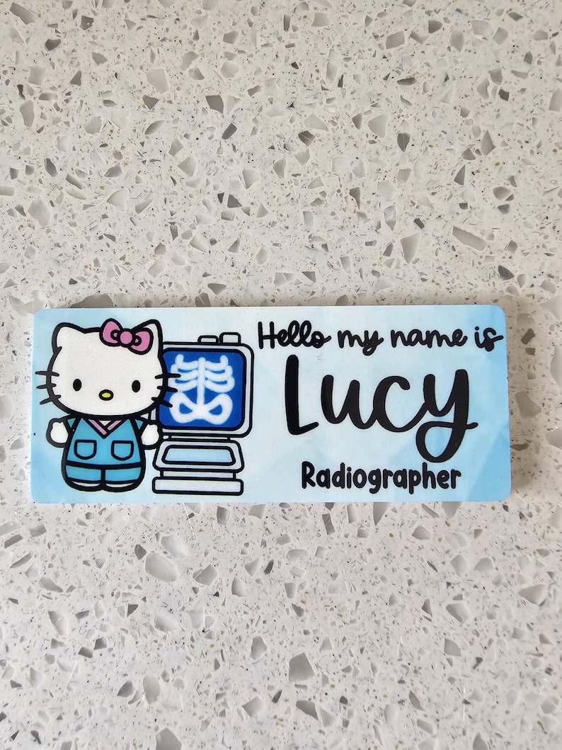 May include: A rectangular name badge with a light blue background featuring a cartoon cat wearing blue scrubs next to an X-ray machine. The text reads Hello my name is Lucy Radiographer in black script and sans-serif fonts.