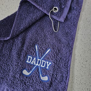 May include: Purple golf towel with a white embroidered design of crossed golf clubs and the word "DADDY".