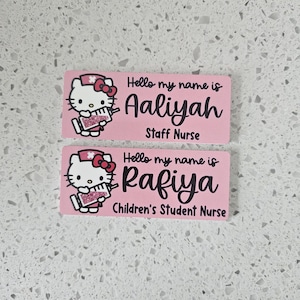May include: Two rectangular pink name badges featuring a cartoon cat dressed as a nurse holding a syringe. The top badge reads Hello my name is Aaliyah Staff Nurse. The bottom badge reads Hello my name is Rafiya Children's Student Nurse.