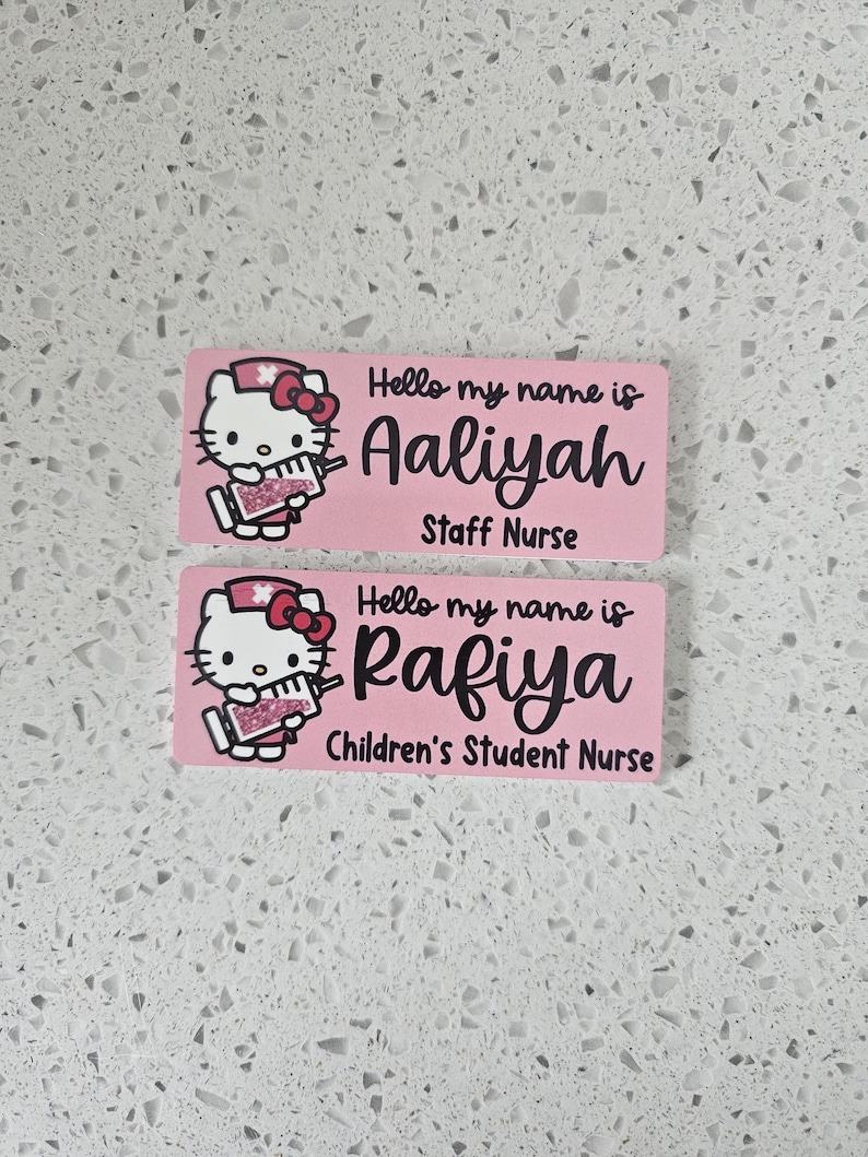 May include: Two rectangular pink name badges featuring a cartoon cat dressed as a nurse holding a syringe. The top badge reads Hello my name is Aaliyah Staff Nurse. The bottom badge reads Hello my name is Rafiya Children's Student Nurse.