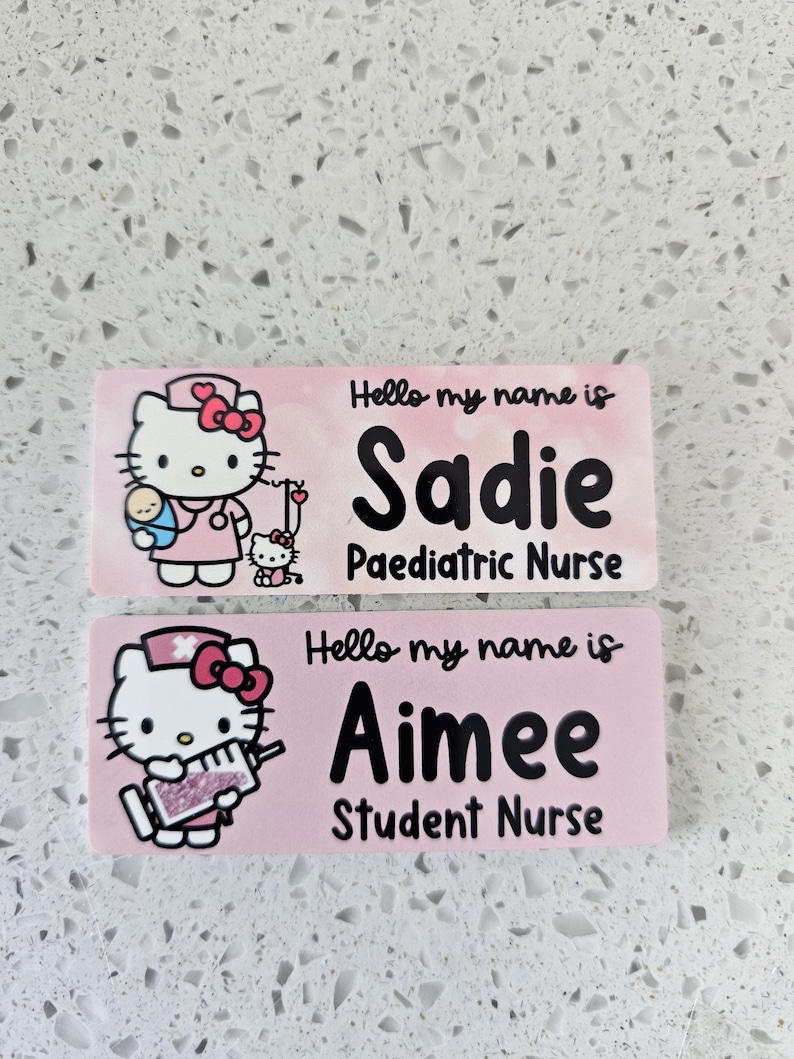 May include: Two rectangular pink name badges featuring Hello Kitty illustrations. The top badge reads Hello my name is Sadie Paediatric Nurse with a nurse Hello Kitty holding a baby. The bottom badge reads Hello my name is Aimee Student Nurse with a nurse Hello Kitty holding a syringe.