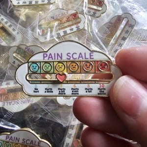 May include: A cloud-shaped enamel pin featuring a pain scale with six coloured faces ranging from blue to red. A sliding pink heart marker indicates pain levels from 0 to 10, with text labels including No hurt, Hurts a little, Hurts a bit more, and Hurts the worst.