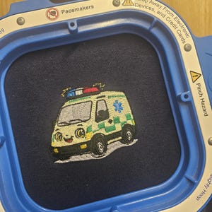 May include: Embroidered ambulance patch with green and yellow details on navy fabric, held in a blue embroidery hoop. The hoop has text including "Pacemakers" and "Keep Away From Electronic Devices, and Credit Cards."