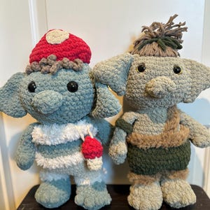 May include: Two handmade crochet dolls. One doll is light blue with a red mushroom hat and a red ice cream cone. The other doll is gray with a green and brown outfit and a brown hair. Both dolls have large black eyes.