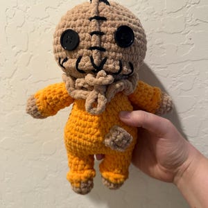 May include: A handmade crochet doll featuring a tan burlap-textured head with black button eyes and stitched facial details. The doll wears a bright orange jumpsuit with a small tan patch on the left leg and a tan textured collar around the neck.