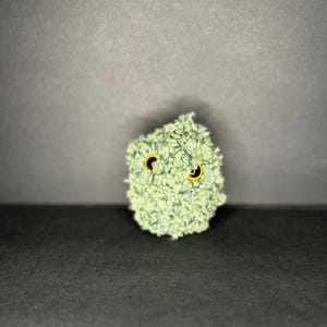 Pode incluir: Uma criatura de pelúcia verde claro e texturizada com olhos amarelos e pretos. O item feito à mão está sobre um fundo cinza escuro. O brinquedo de pelúcia tem um design único e caprichoso.