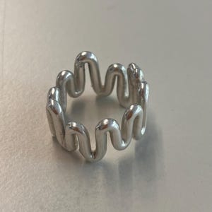 Könnte beinhalten: Ein silberner Ring mit einem wellenförmigen, organischen Design. Der Ring ist aus glattem, poliertem Metall gefertigt und hat einen einzigartigen, modernen Look.
