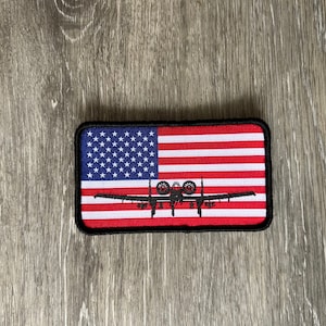 May include: A black and white embroidered patch with an American flag and a silhouette of an A-10 Warthog aircraft.