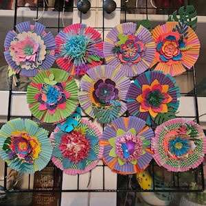 Puede incluir: Una colección de coloridas flores de papel hechas a mano en varios tamaños y diseños. Cada flor presenta pétalos en capas en tonos de rosa, morado, verde y naranja, con detalles intrincados. Las flores se exhiben en una rejilla de alambre negra.