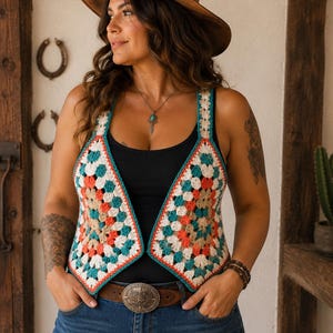 Op de afbeelding: Een mouwloos gehaakt vest met een veelkleurig granny square patroon in tinten wit, groenblauw en koraal. Het vest heeft een V-hals en wordt gedragen over een zwart hemdje, gecombineerd met een blauwe spijkerbroek en een bruine leren riem.